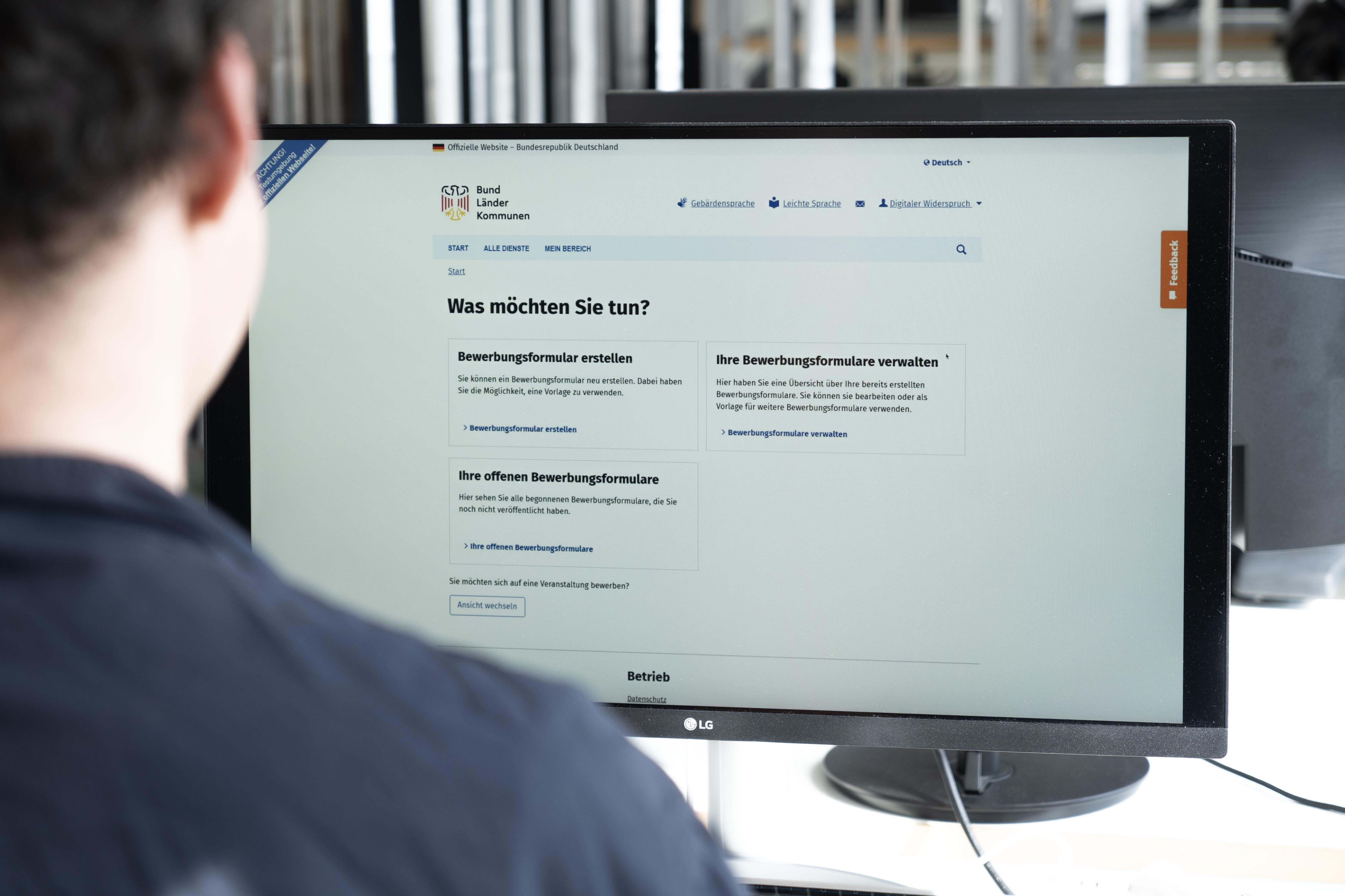 Person vor einem Computermonitor, der ein Eingabesystem zeigt, auf dem Bewerbungsformulare erstellt, verwaltet und bearbeitet werden können; eine offizielle Website der Bundesrepublik Deutschland mit einem schwarz-rot-goldenem Adler beschriftet Bund-Länder-Kommunen