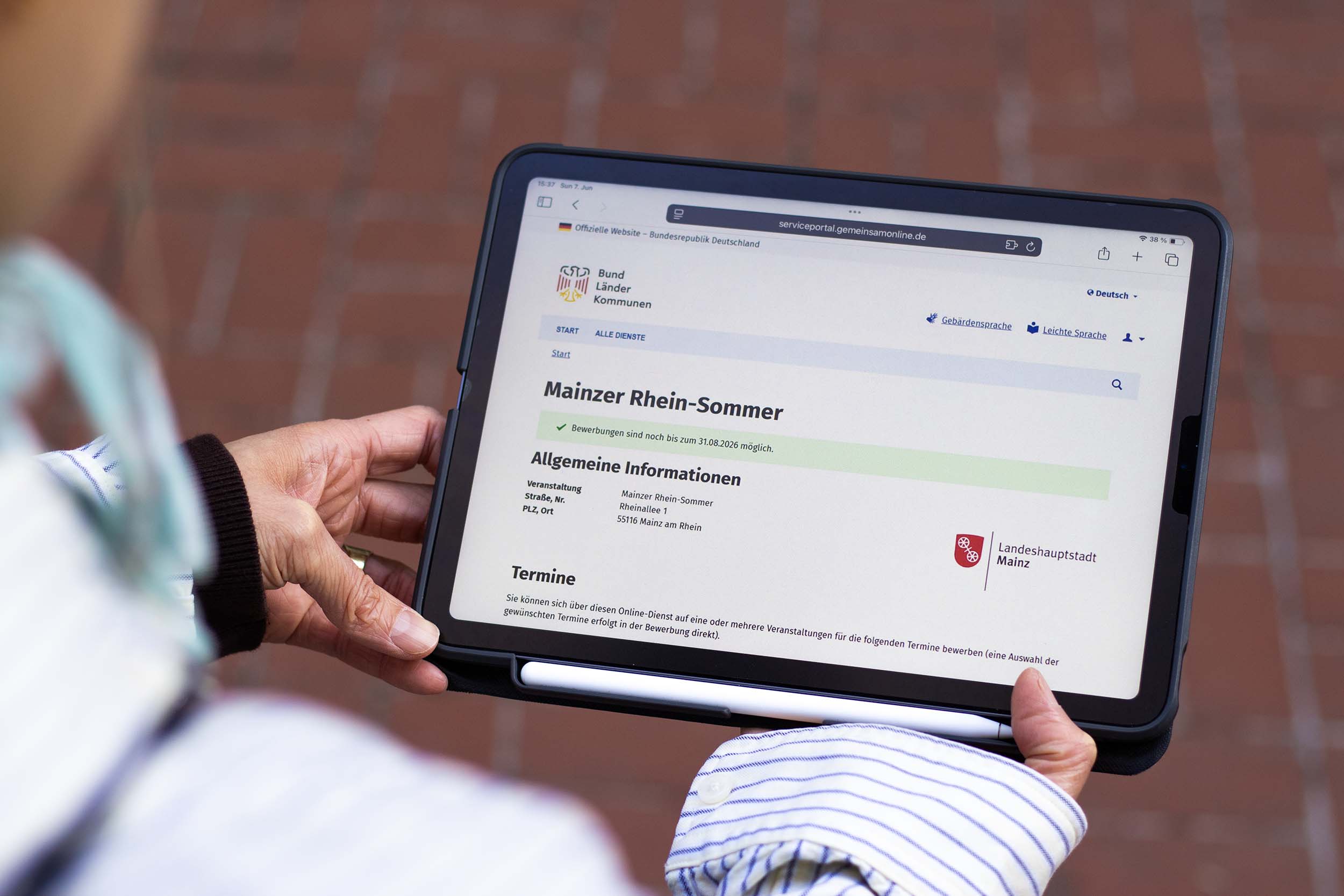 Person mit Tablet in der Hand, auf dem der Bewerbungsprozess für den Mainzer Rhein-Sommer zu sehen ist; eine offizielle Website der Bundesrepublik Deutschland mit einem schwarz-rot-goldenem Adler beschriftet Bund-Länder-Kommunen