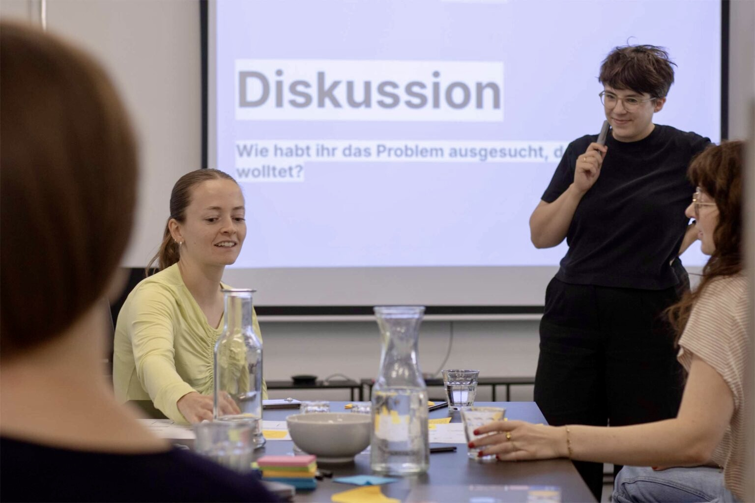 4 Frauen mit heller Haut in einem Workshop-Setting, hinter ihnen eine Leinwand mit der Projektion „Diskussion – Wie habt ihr das Problem ausgesucht, das ihr lösen wolltet?'"